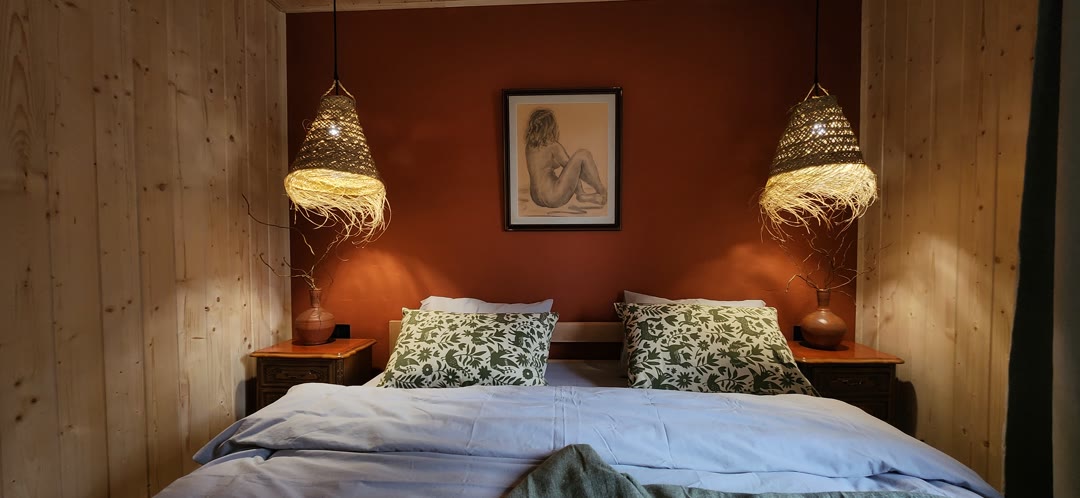 The bedroom with a terracotta wall, two woven lamps and a drawing above the bed