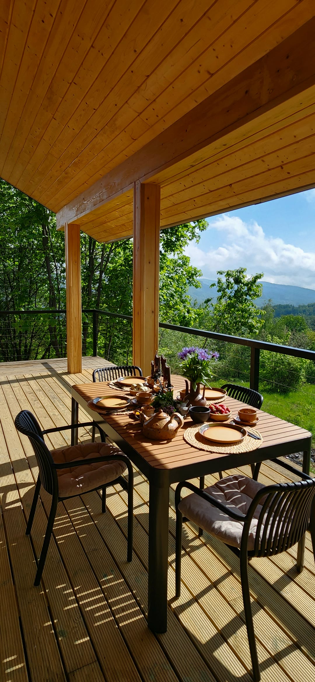 A breakfast table on the terrace under the timber roof, forest and mountains beyond